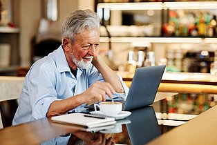 Ein Mann im mittleren Lebensalter blickt in eine Laptop und rührt zugleich in einer Tasse Kaffee.